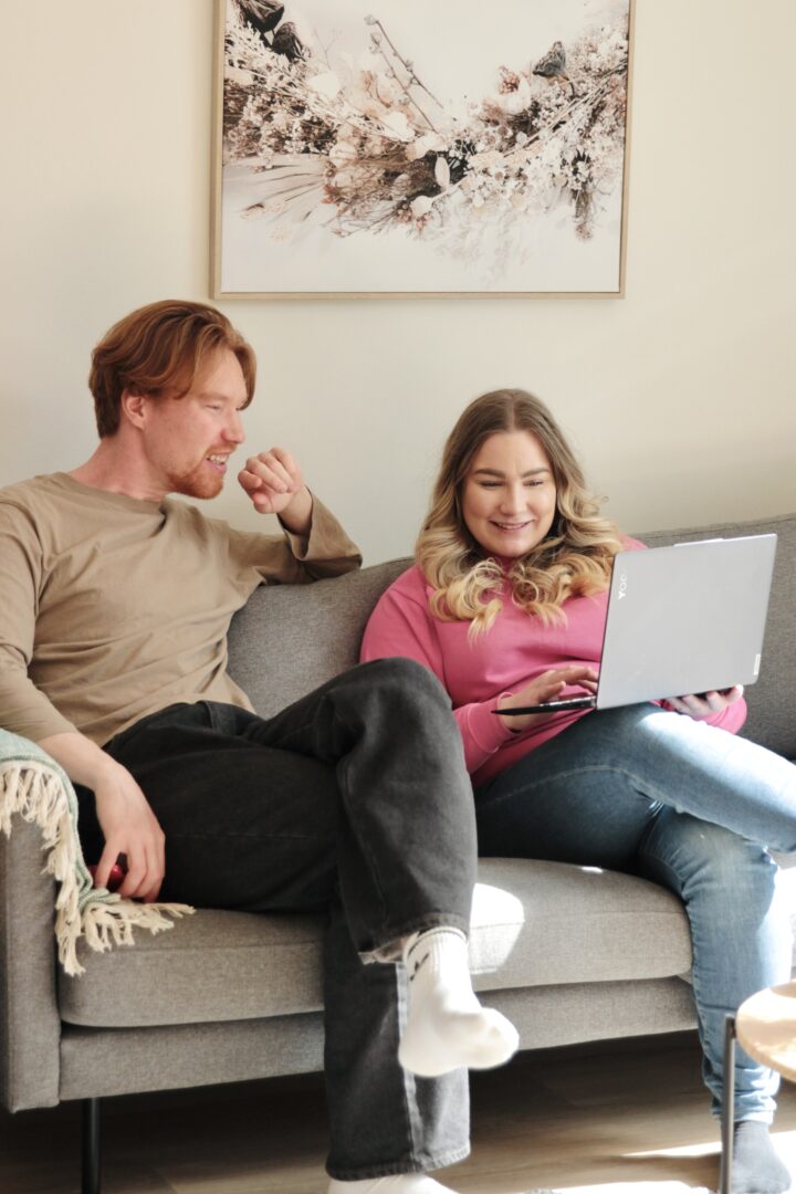 Two people sit on a gray sofa; man on the left looks toward the woman, who is using a silver laptop on her lap in a living room.