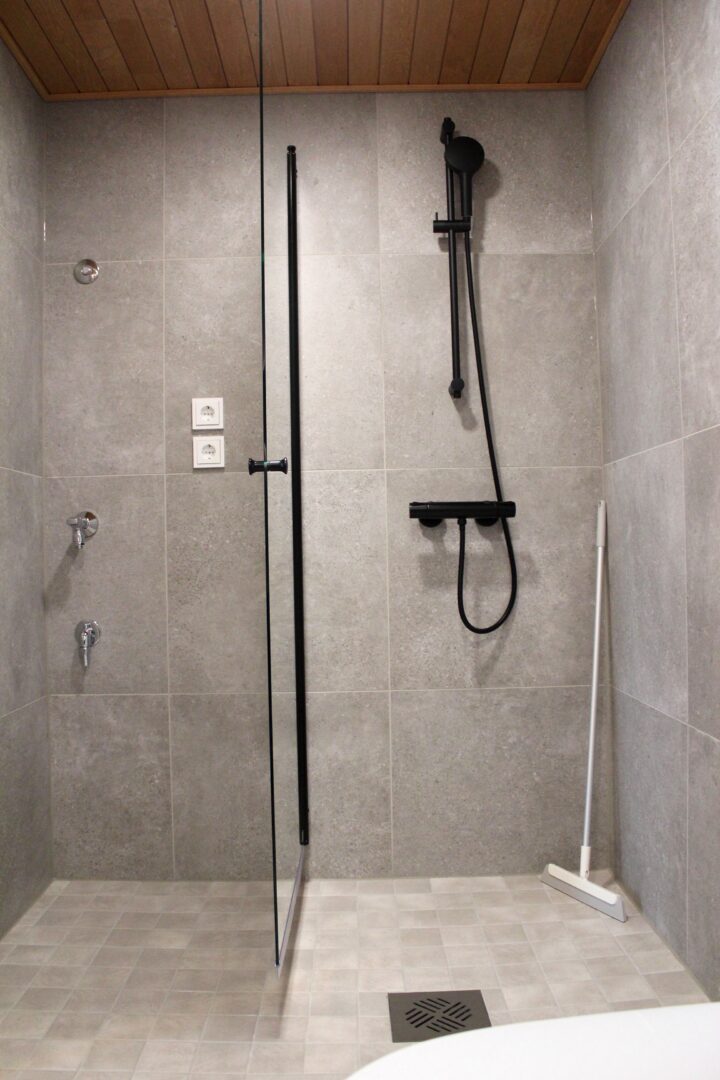 Gray tiled walk-in shower with a glass divider and black handheld shower fixtures on a dark rail set, plus a floor drain nearby.