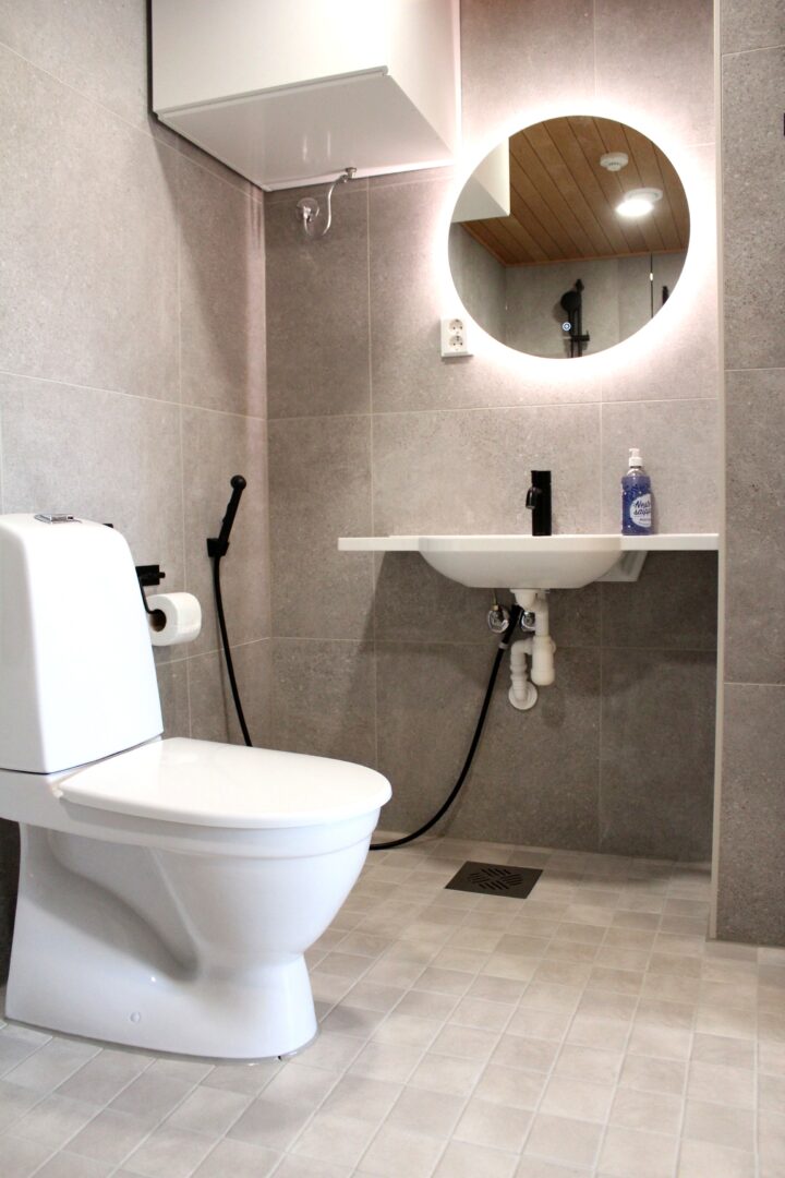 Modern gray-tiled bathroom with a white wall-mounted sink, round backlit mirror, and black faucet; toilet on the left and a handheld bidet sprayer nearby, soap bottle on the sink, tiled floor and wall drain visible.