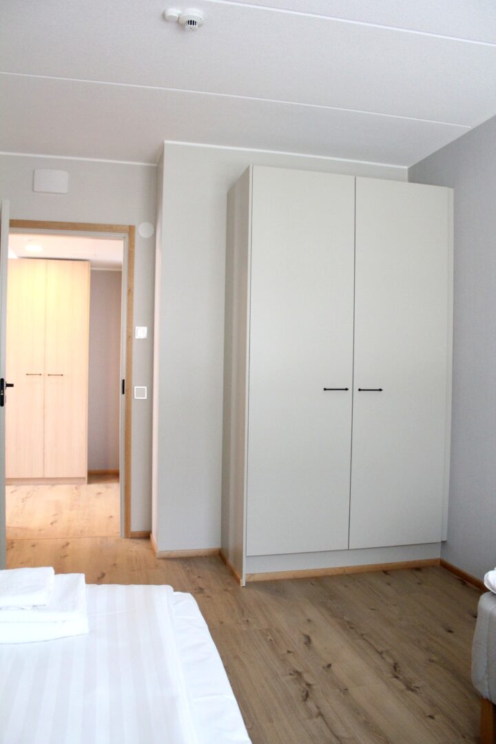 Bedroom with light gray walls, a tall white wardrobe, and a wooden floor; bed visible in the foreground left.