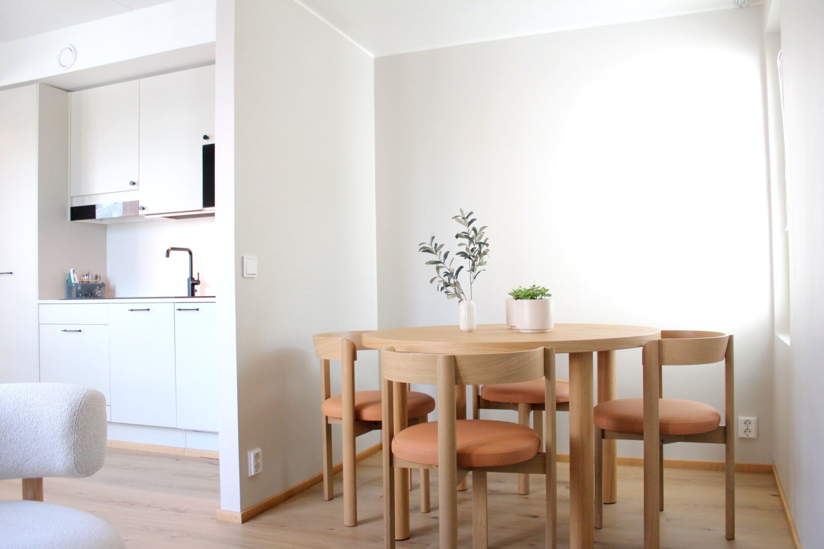 Bright dining area with a round wooden table and matching stools, small plants on the table, and white kitchen cabinets along the left wall.