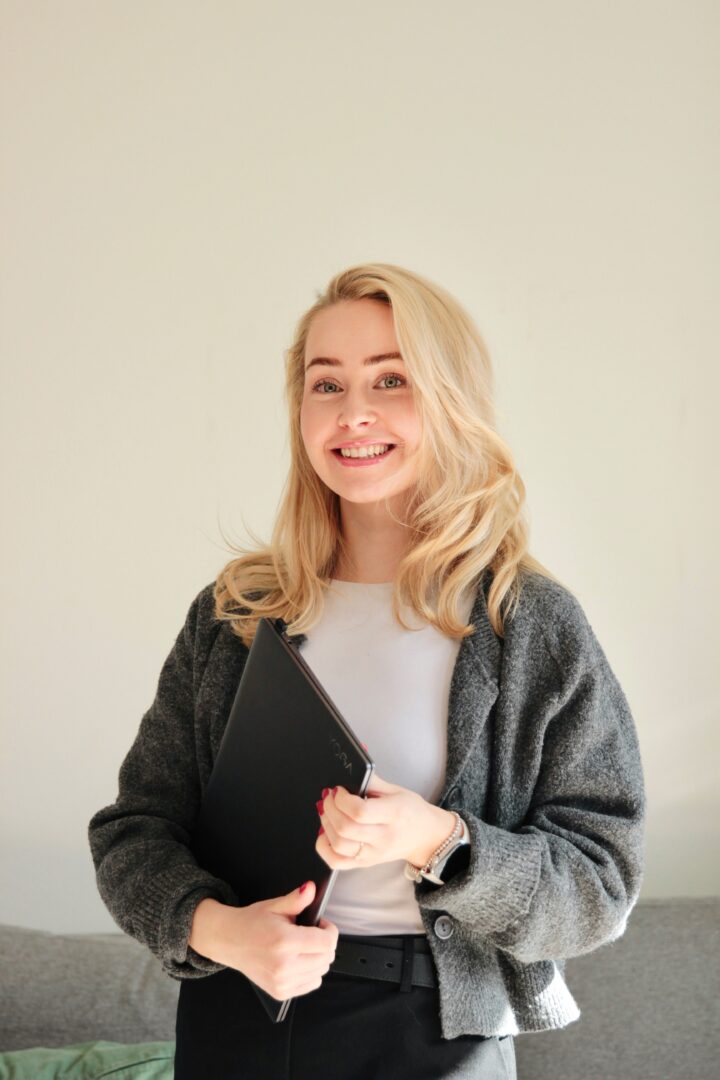 Smiling woman with long blond hair holding a black notebook, wearing a gray cardigan and white top against a light background.