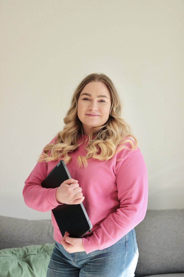 Woman standing indoors in a pink sweater, holding a black notebook and smiling at the camera.